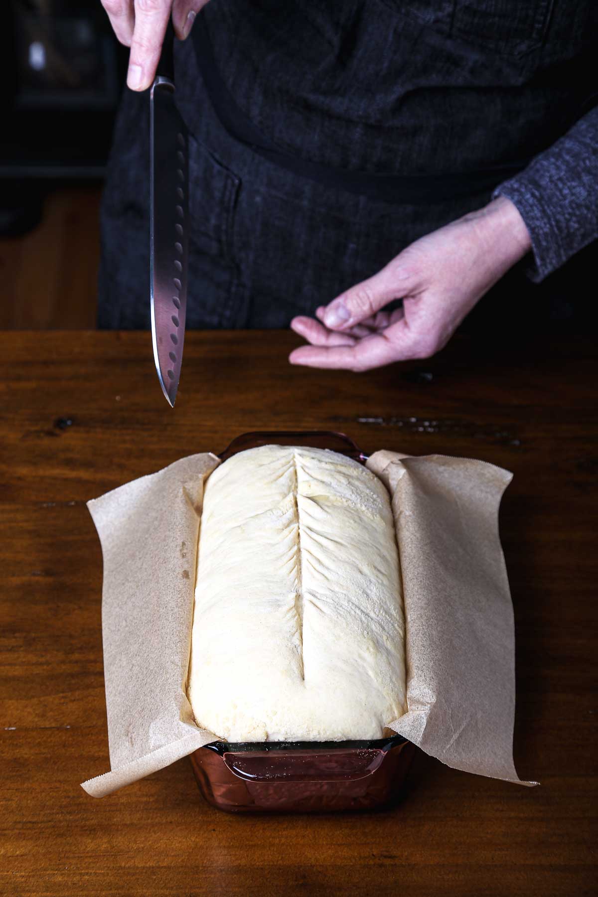 Loaf dough in a parchment-lined pan being scored down the center with a knife before baking.