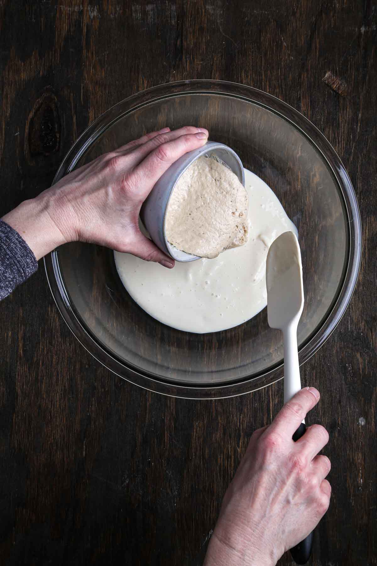 Bloomed yeast being poured into a bowl of blended tofu mixture with a spatula nearby.