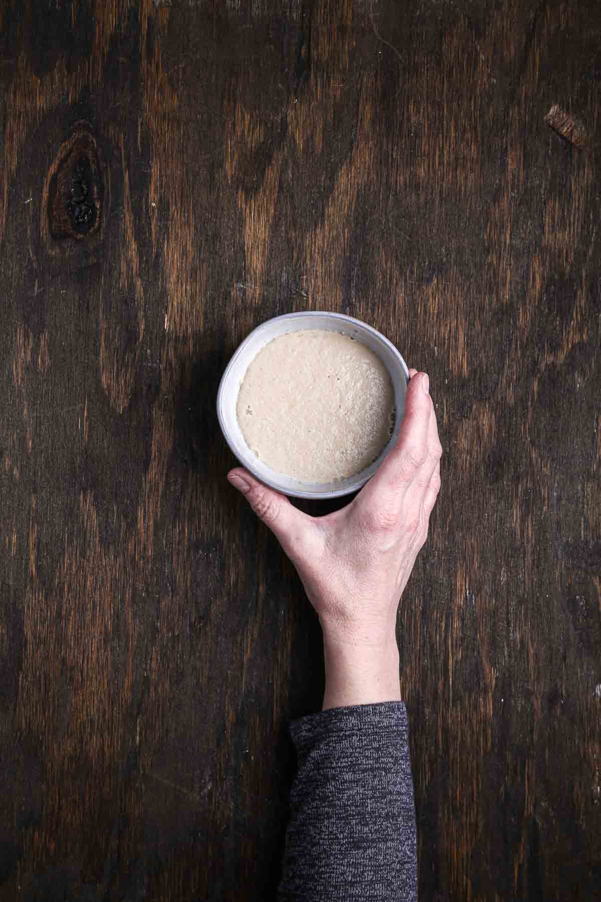 Fully proofed yeast forming a creamy, bubbly foam in a small bowl held over a dark wood surface.