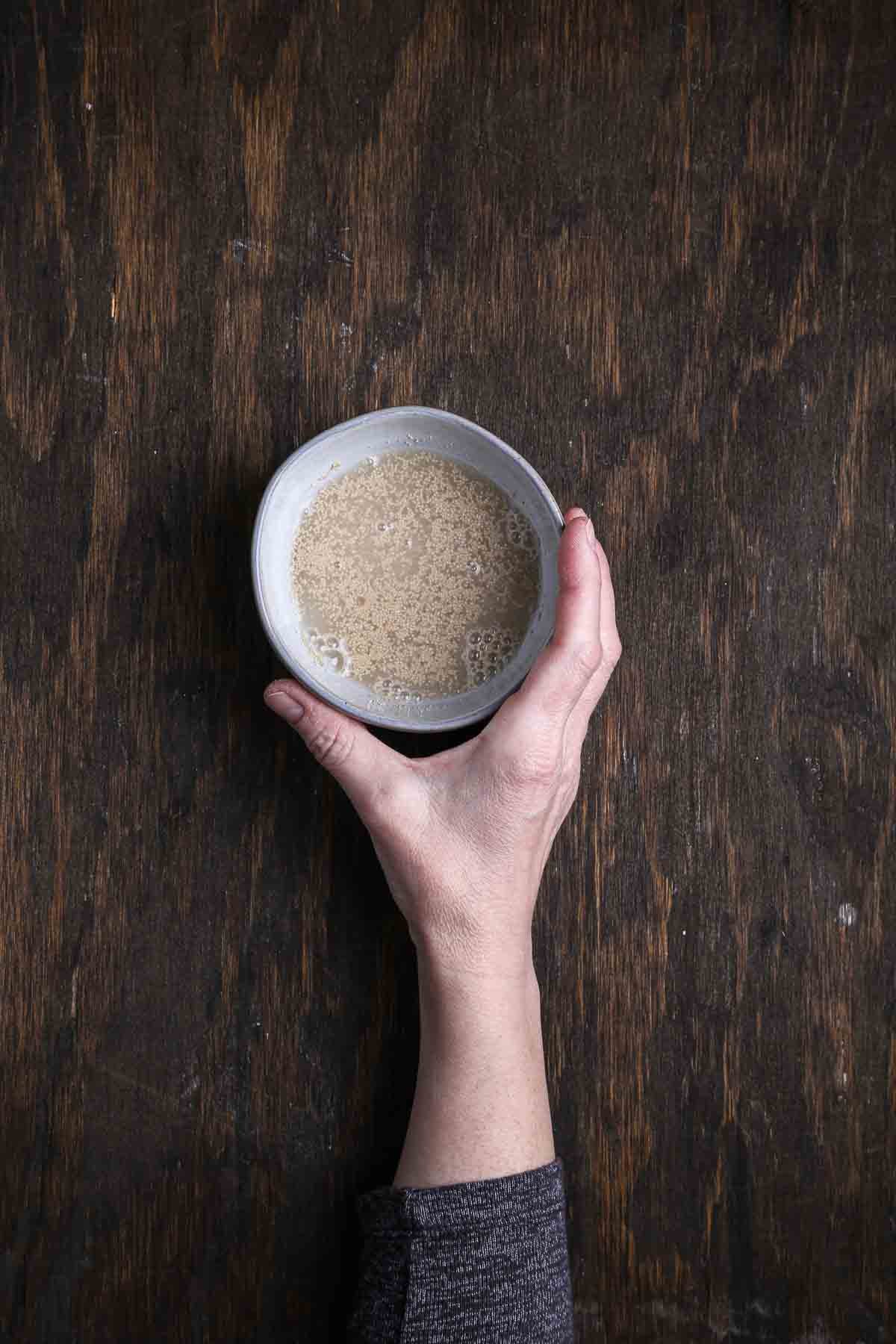 Yeast granules bubbling in warm water during a proofing test, held in a small bowl over a dark wood surface.