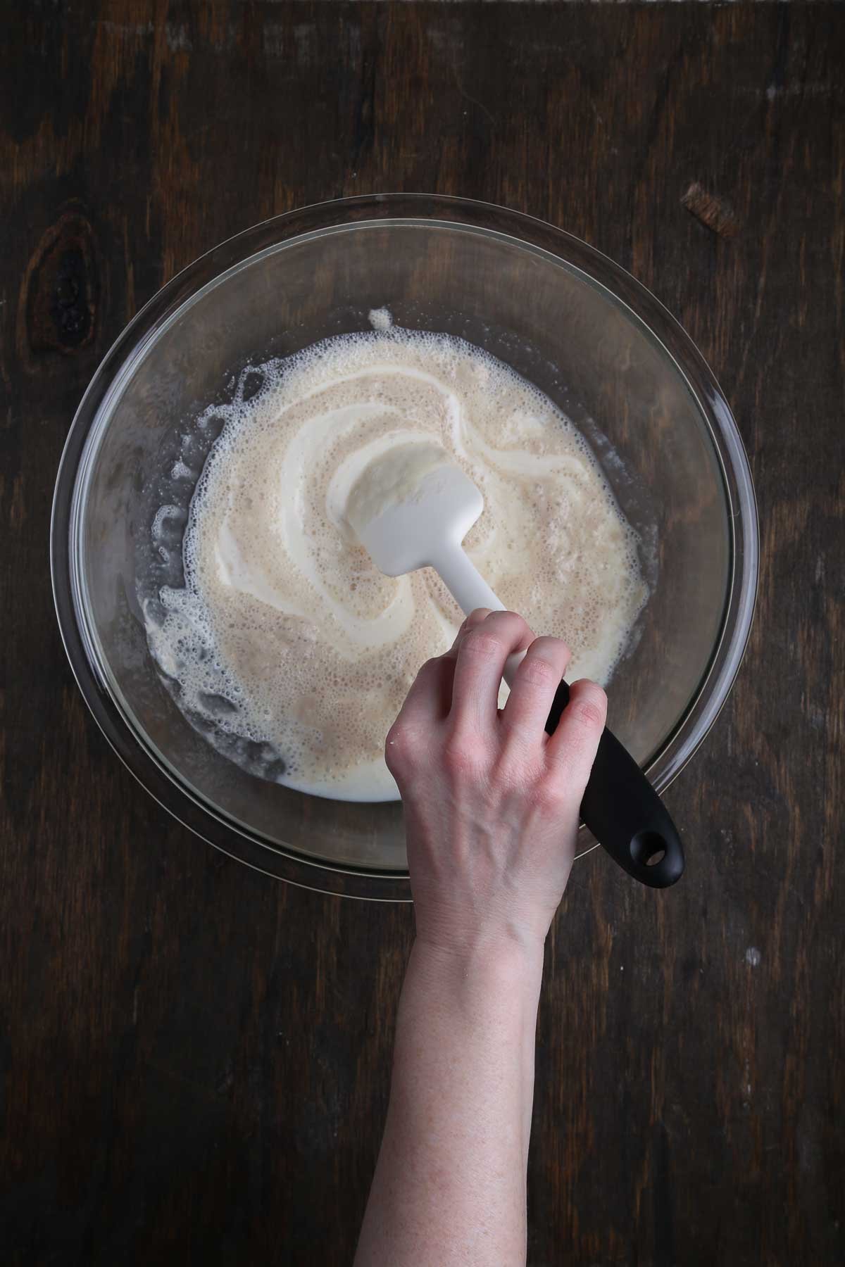 A hand stirring the combined tofu and yeast mixture in a large glass mixing bowl.