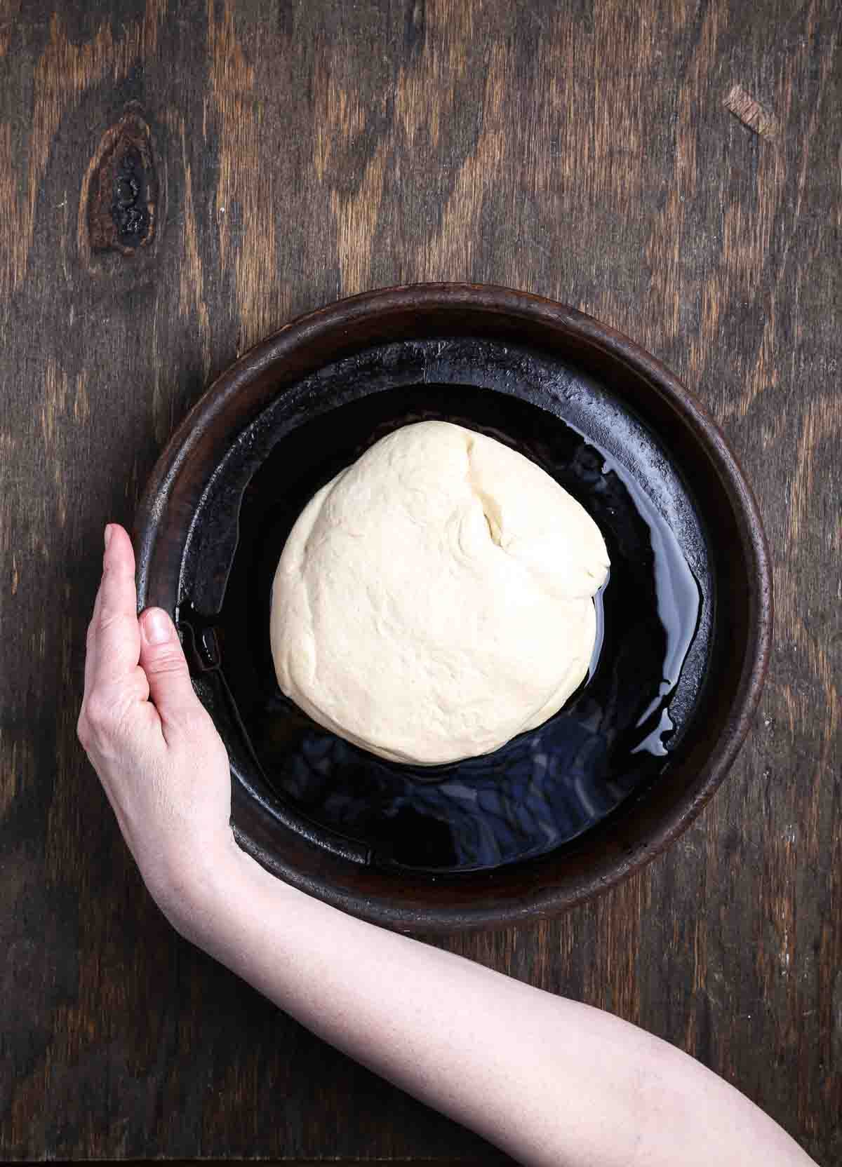 Ball of pizza dough resting in olive oil inside a round baking pan before proofing.