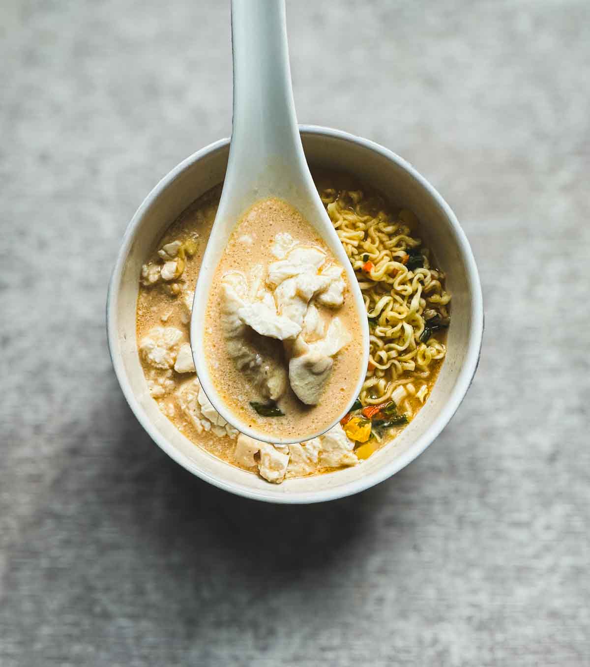 Soft tofu cubes resting in a creamy broth, lifted on a spoon from a bowl of ramen-style soup with noodles and vegetables.